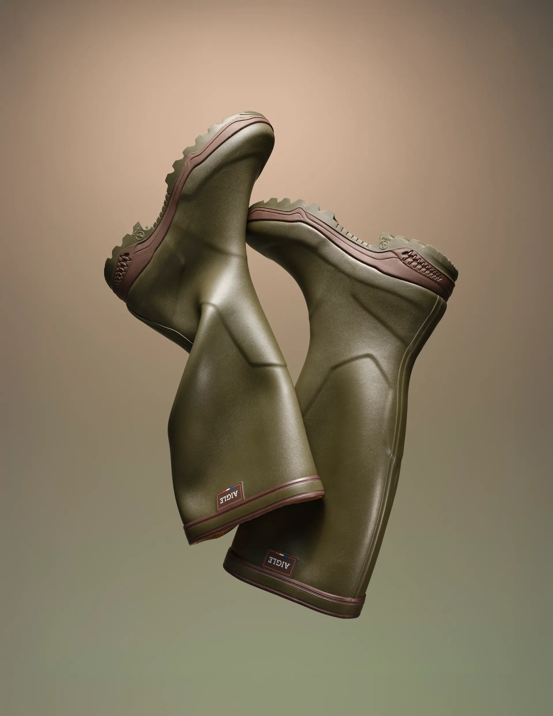 Une paire de bottes en caoutchouc vert olive avec des semelles marron, suspendues sur un fond dégradé beige et vert.