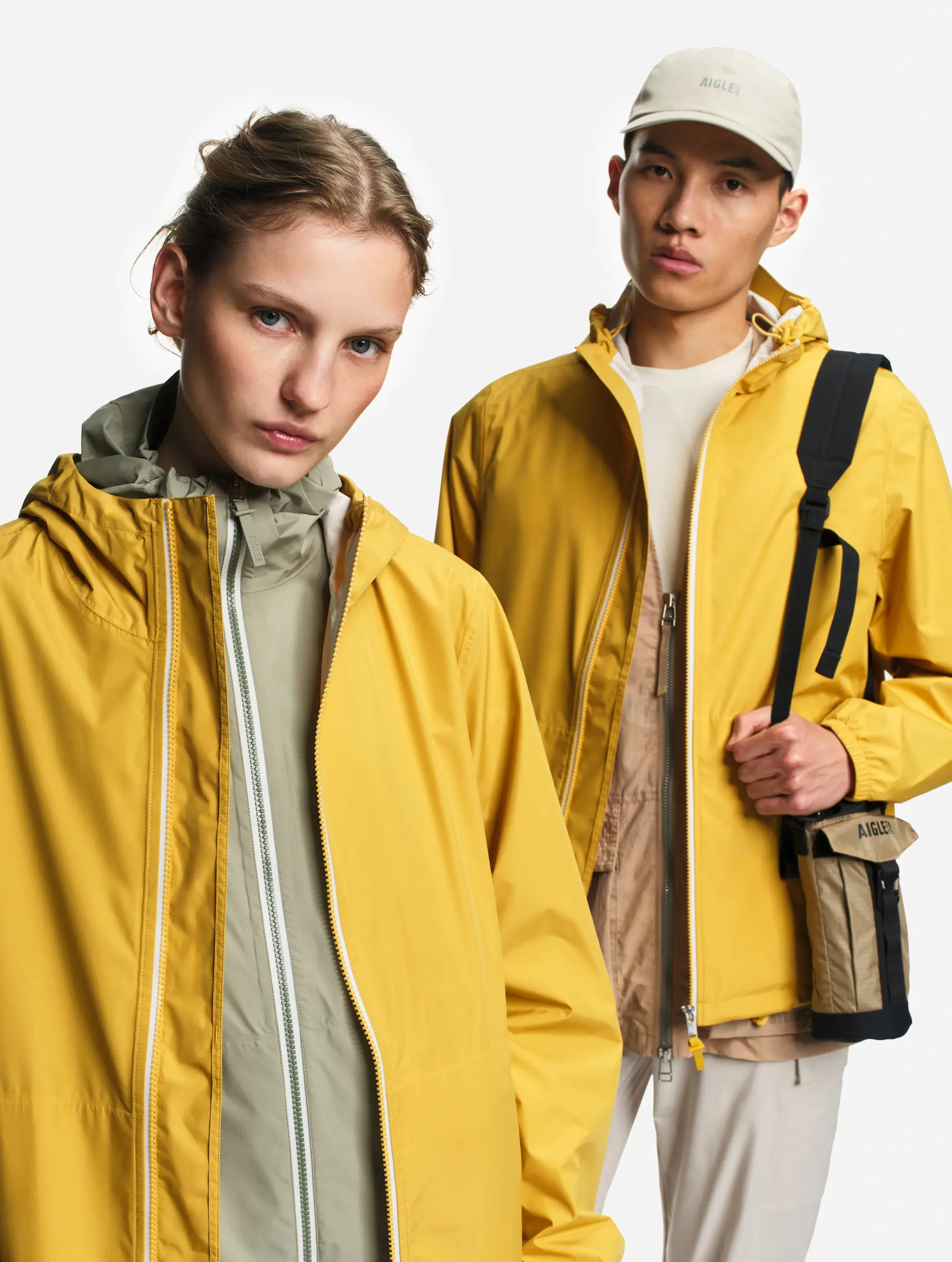 Portrait de deux modèles portant des vestes jaunes imperméables, la femme au premier plan et l'homme en arrière-plan avec une casquette et un sac bandoulière.