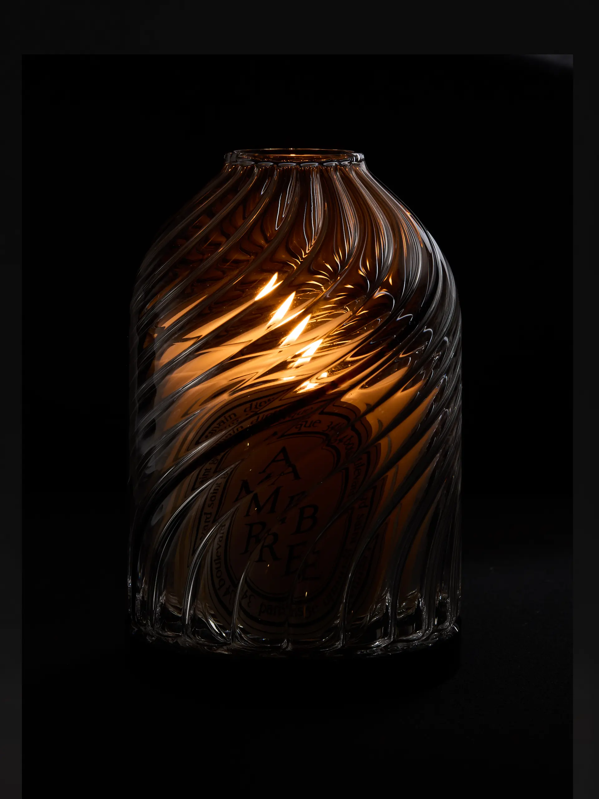 Bougie allumée dans un pot en verre ambré et torsadé avec l’étiquette Ambre visible à l’intérieur.