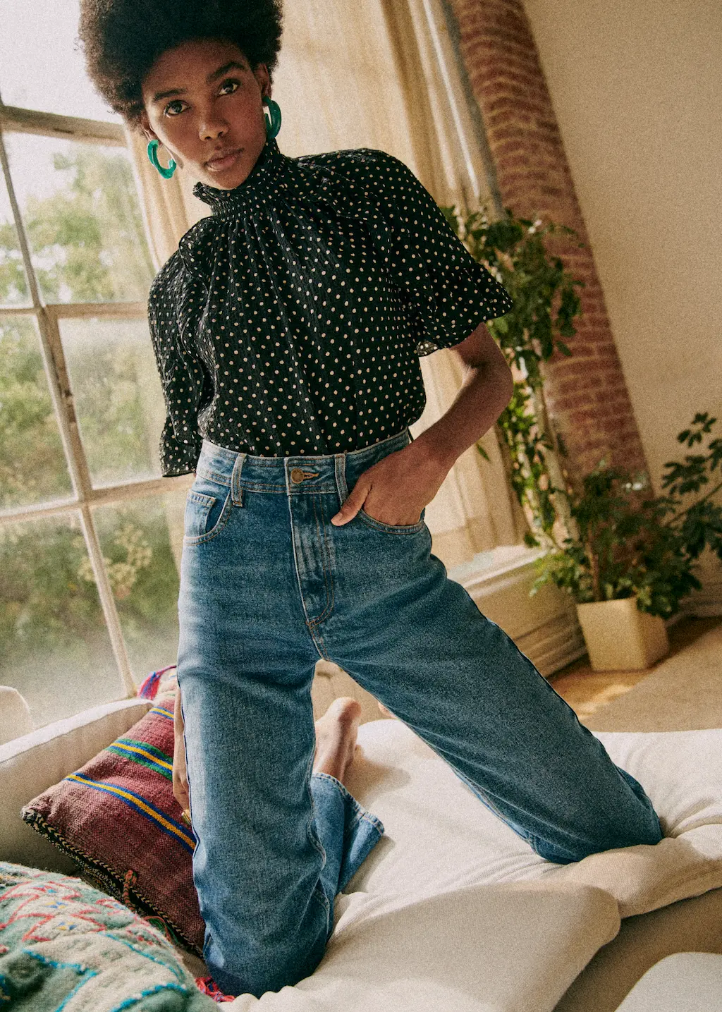 Femme aux cheveux courts bouclés portant un chemisier noir à pois blanc et un jean bleu, posant sur un canapé avec un grand coussin coloré, dans un salon lumineux avec plantes et grande fenêtre.