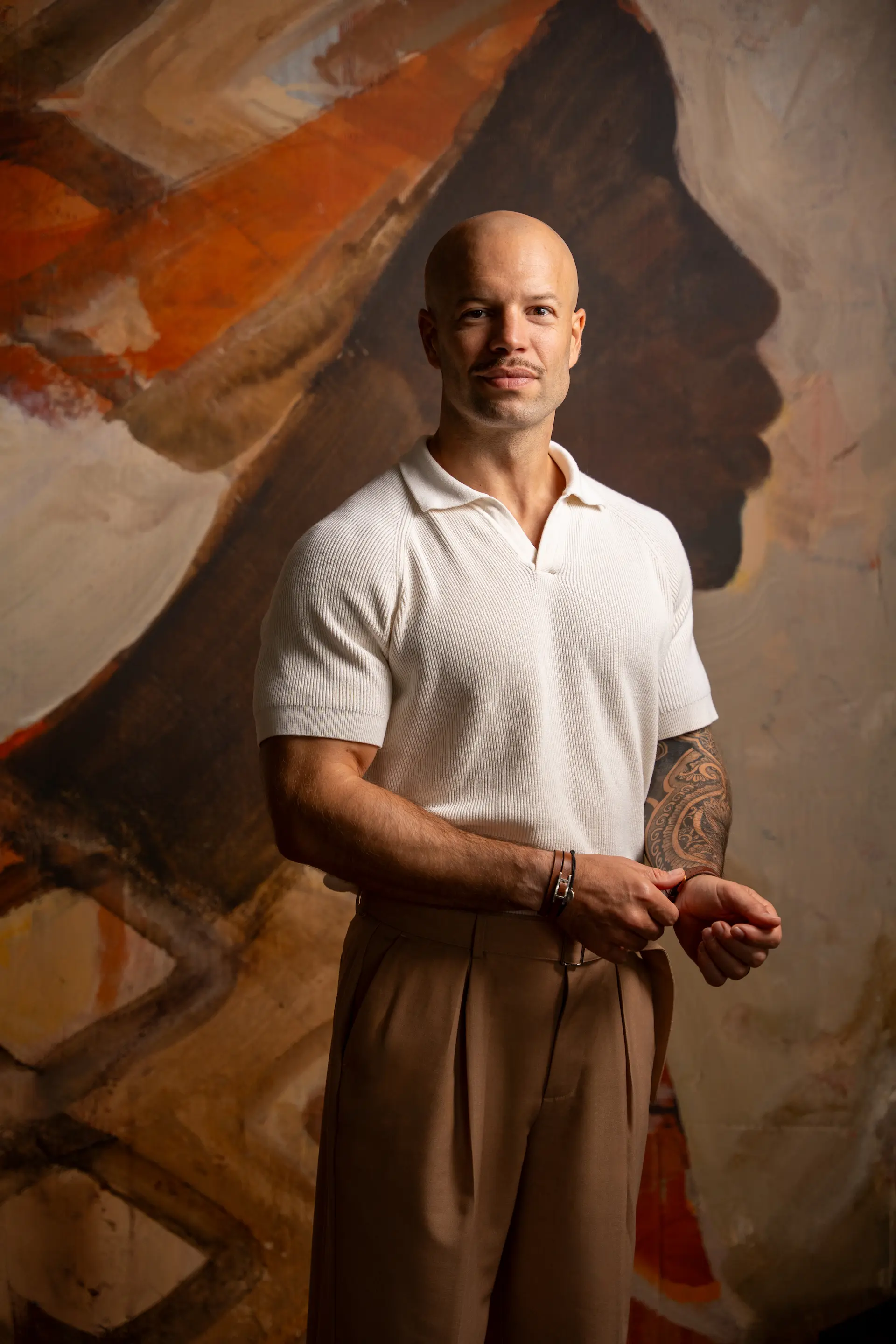 Homme chauve portant un polo blanc et un pantalon beige, posant devant une grande peinture murale avec un profil sombre stylisé.