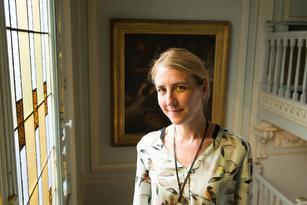 Smiling woman with blonde hair wearing a bird-patterned blouse standing indoors near stained glass window and ornate framed painting.
