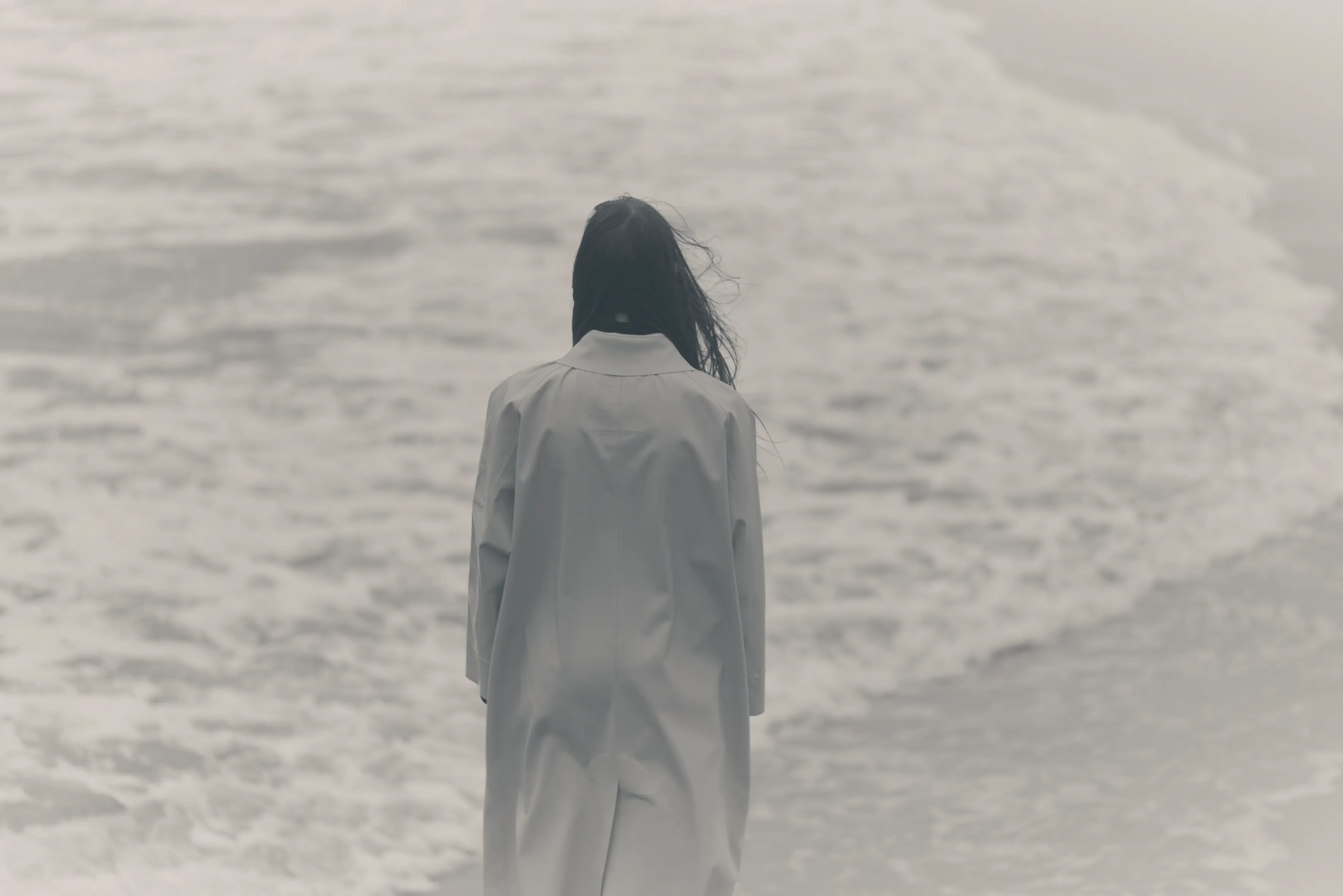 Personne vue de dos portant un manteau clair, regardant la mer agitée avec des vagues.