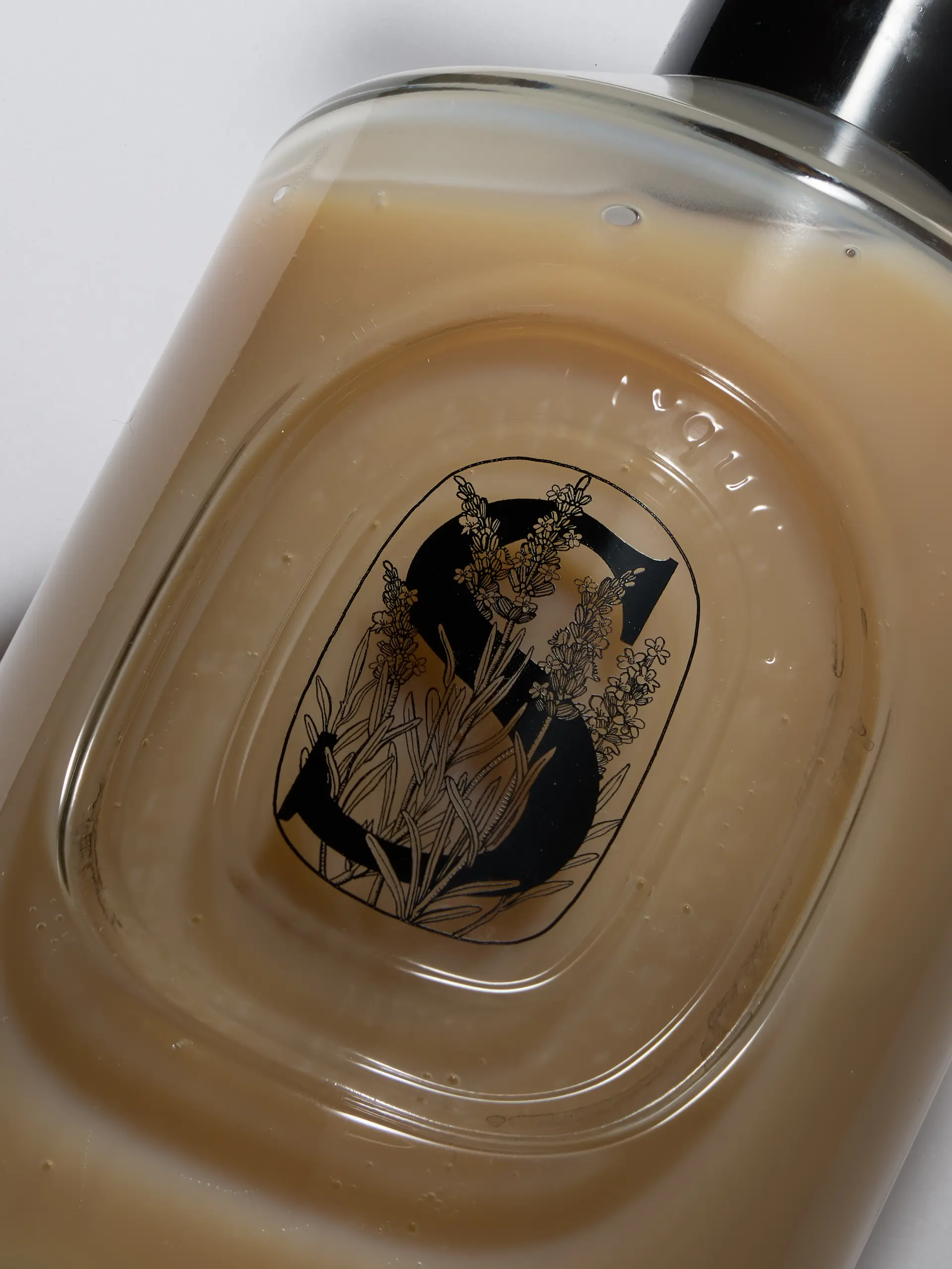 Gros plan sur une bouteille transparente contenant un liquide beige avec une étiquette illustrée d'un grand S et de brins de lavande noire.
