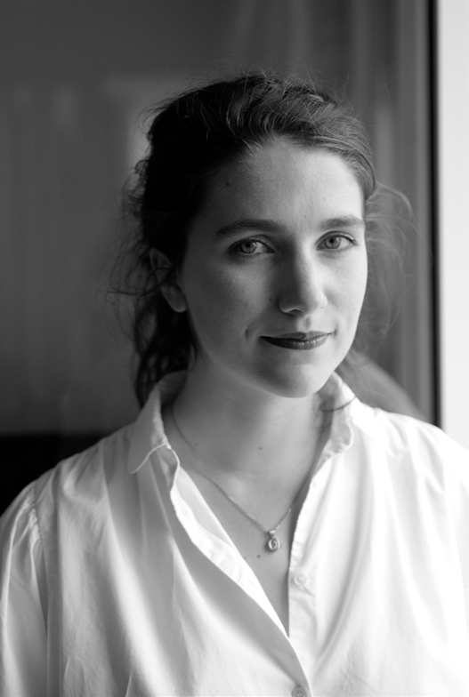 Black and white portrait of a woman with dark hair pulled back, wearing a white shirt and necklace, looking at the camera near a window.
